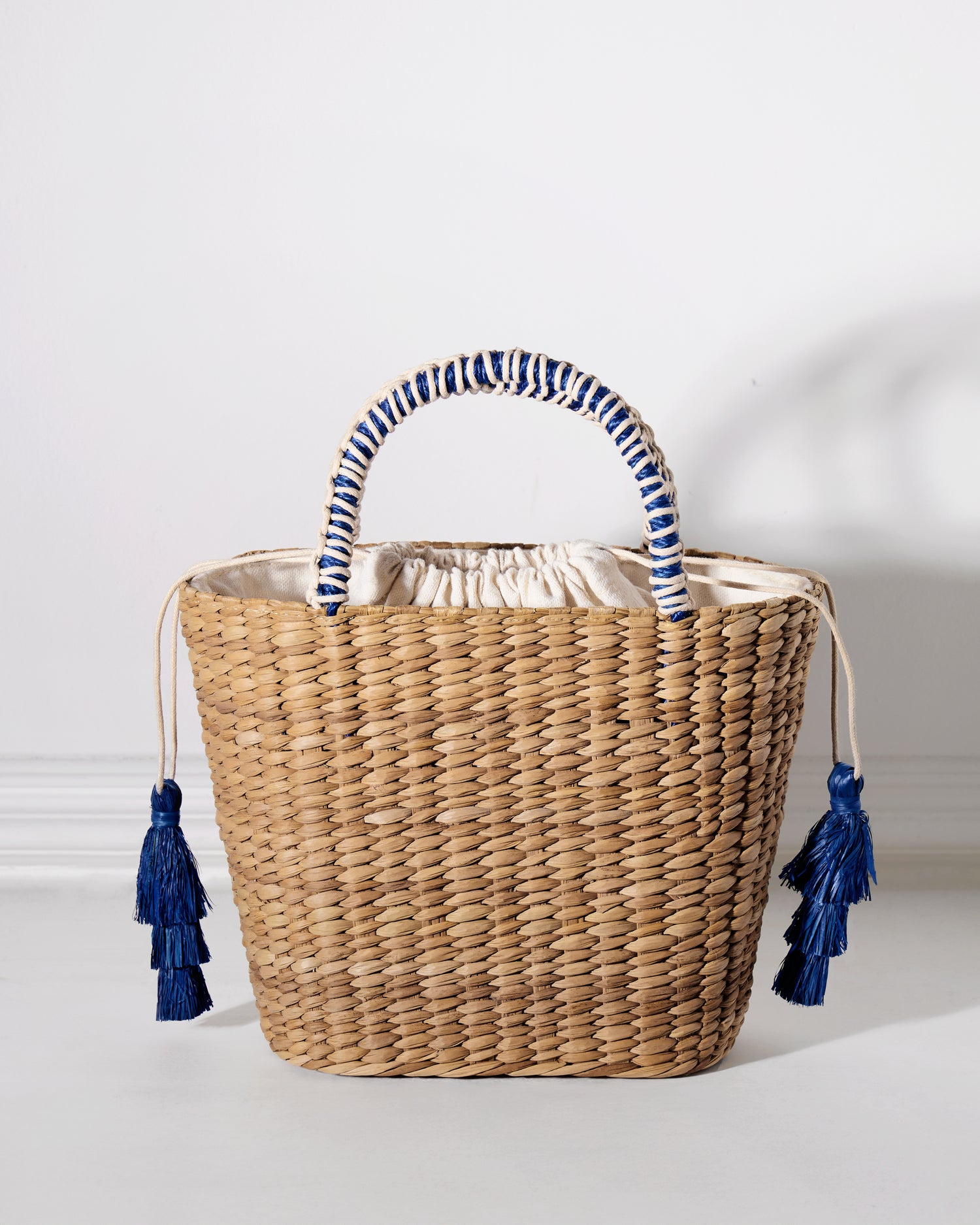 Woven straw basket with a blue and natural-colored cord-wrapped handle, white fabric drawstring closure, and two blue raffia tassels.