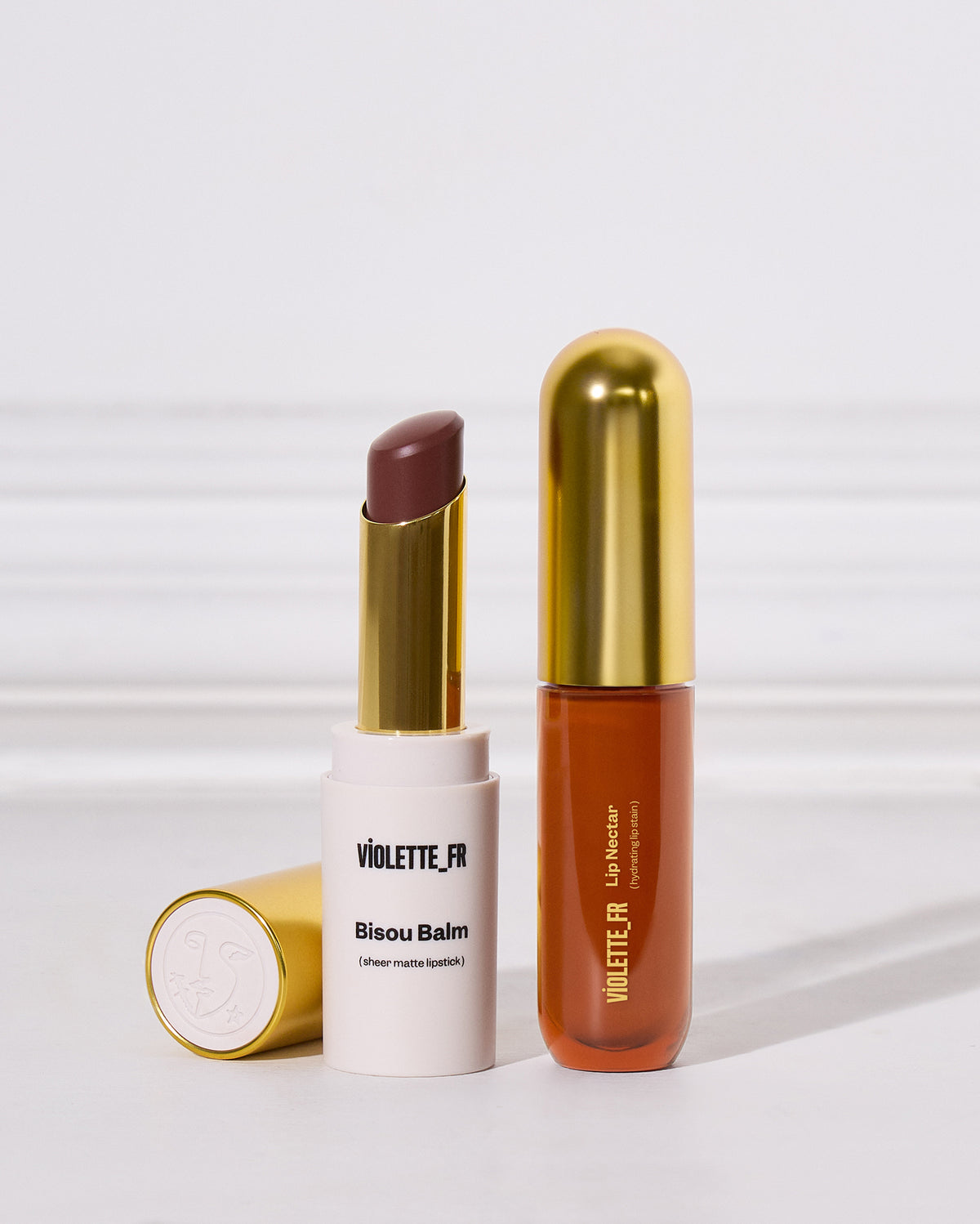 Lip Nectar & Bisou Balm Duo - Two Violette FR cosmetics from the Bisou Balm line, including a white lipstick tube with a gold cap showing a matte cool brown lipstick bullet, and a translucent orange-brown lip liner tube with a gold cap. - Image 1 of 17