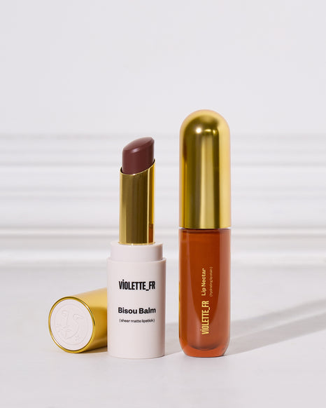 Two Violette FR cosmetics from the Bisou Balm line, including a white lipstick tube with a gold cap showing a matte cool brown lipstick bullet, and a translucent orange-brown lip liner tube with a gold cap.