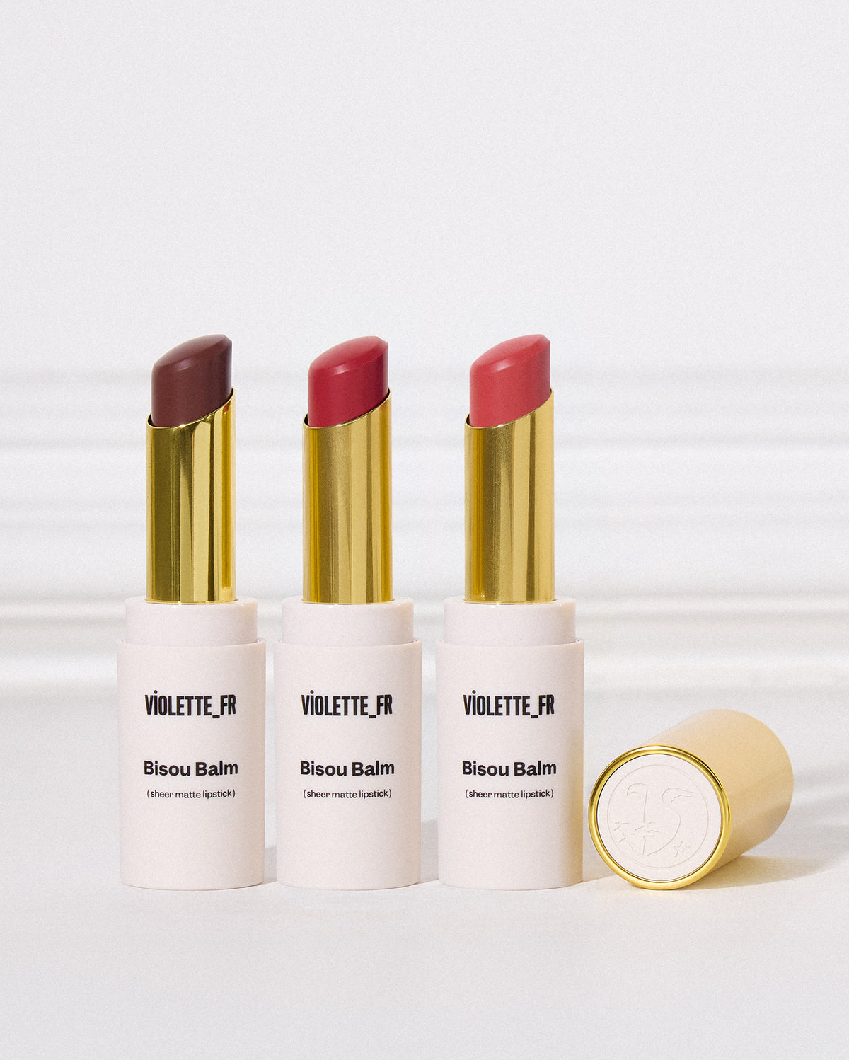 Three Violette.fr Bisou Balm lip balm tubes standing upright on a white surface, with one gold cap placed beside them. The one on the left, a matte cool brown. In the middle, a bright red. On the right, a reddish-pink. - Bisou Balm Trio