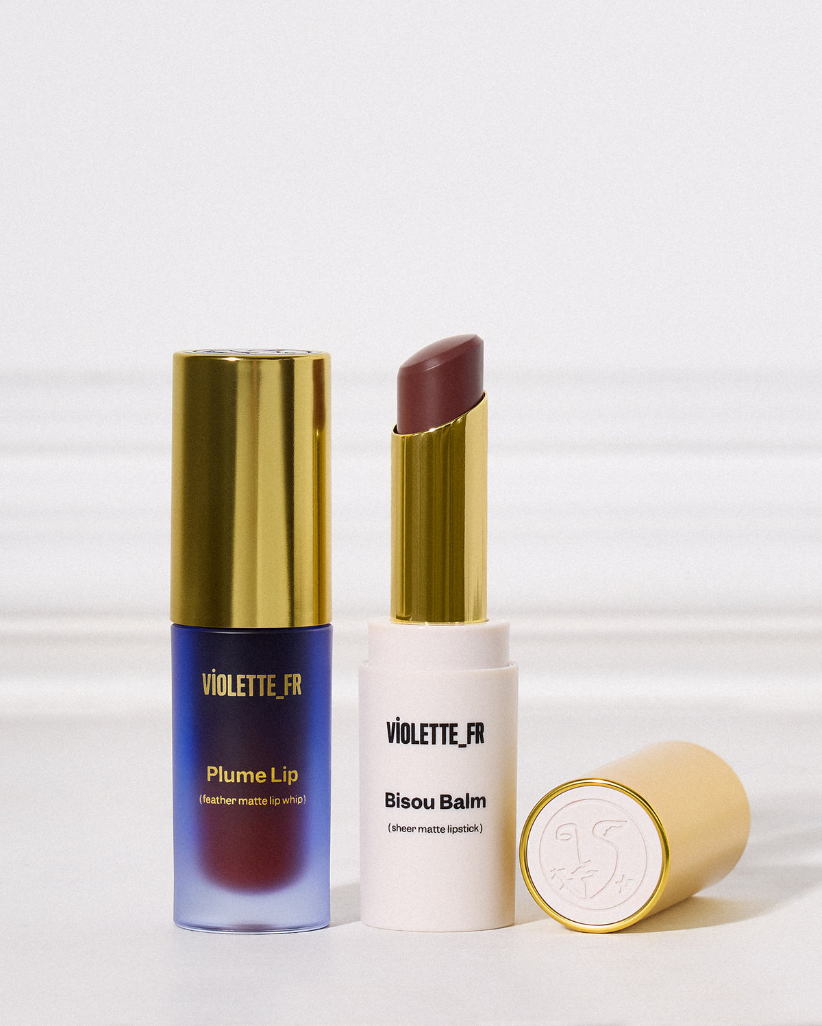 Duo Plume Lip & Bisou Balm - Violette_FR Plume Lip (feather matte lip whip) in a purple-to-red tube and Bisou Balm (sheer matte lipstick) with a rcool brown bullet in a white tube, both with gold accents. - Image 1 of 15