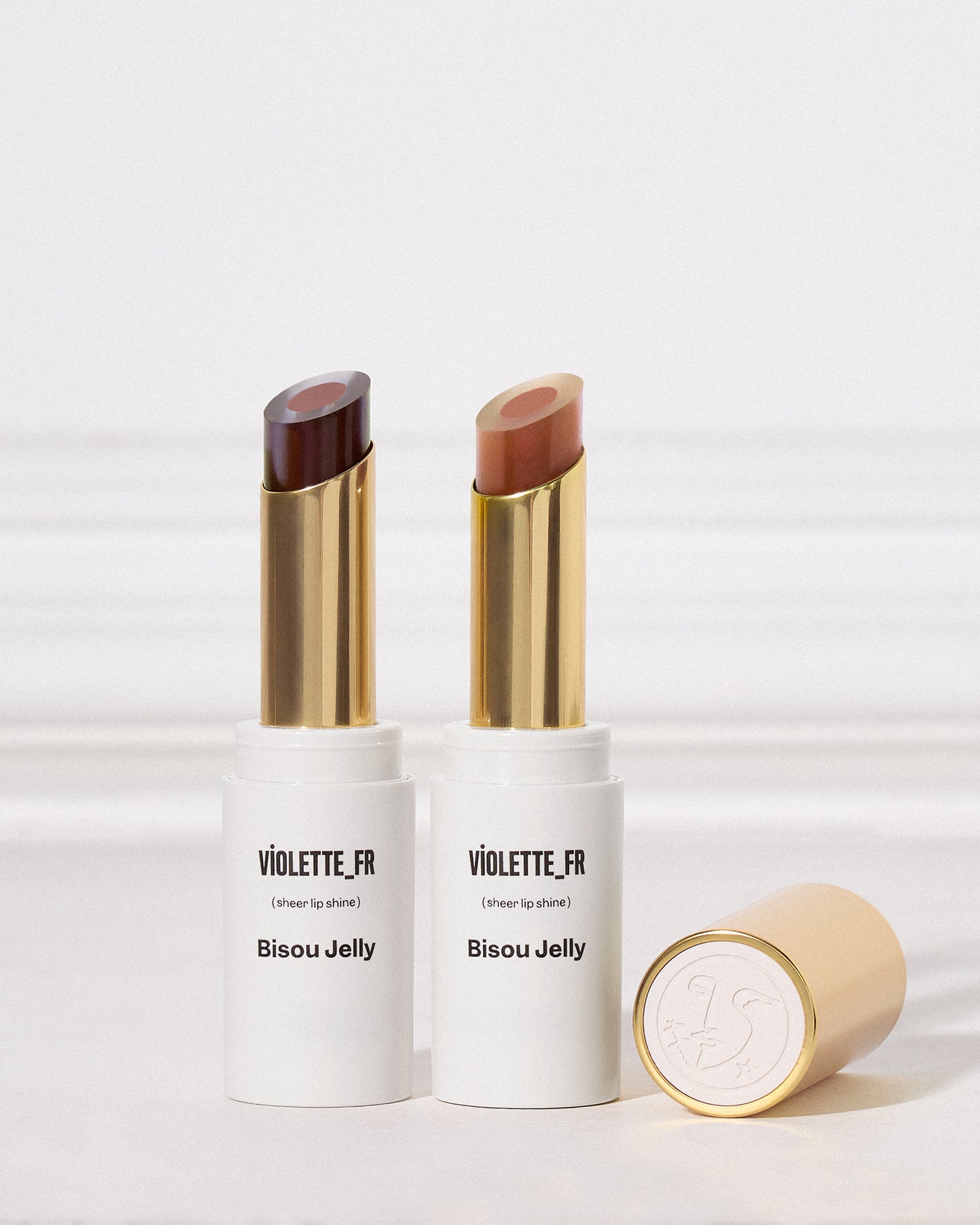 Two lipsticks with sheer outer layers and darker cores stand upright against a white background. The lipsticks have gold and white tubes, with the one on the left featuring a deep purple shade and the one on the right a nude pink shade. A gold cap embossed with a logo lies in the foreground. - Bisou Jelly Duo