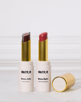 Two lipsticks in white and gold tubes are standing upright next to one another on a white surface. The left is a two-toned sheer purple shade, and the right is a matte red. A cap rests nearby.