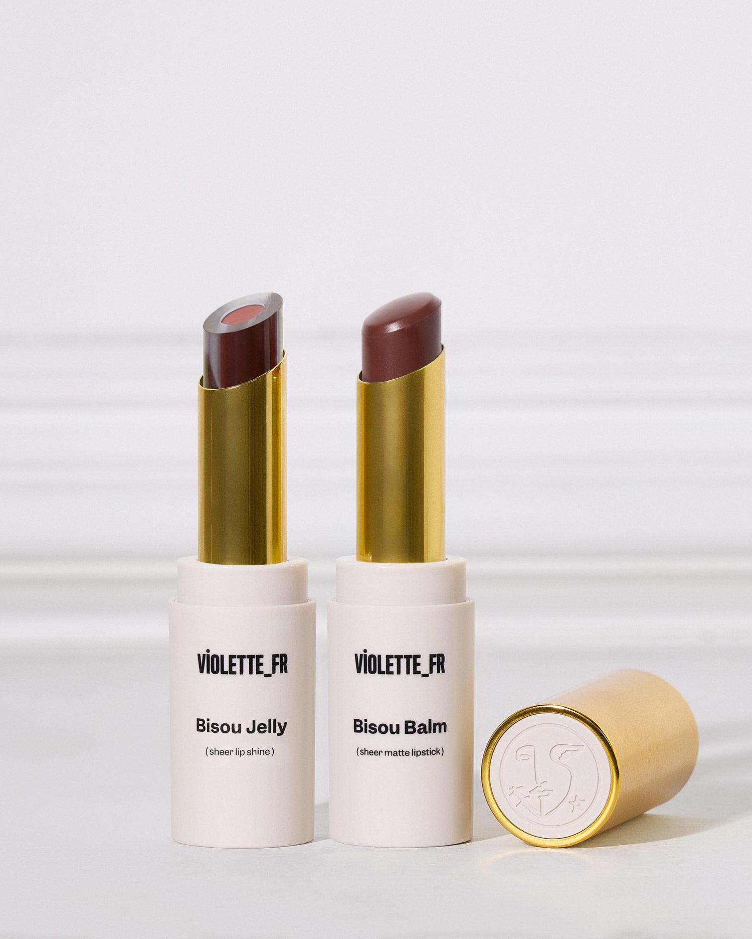 Bisou-Bisou Lip Duo - Two lipsticks in white and gold tubes are standing upright next to one another on a white surface. The left is a two-toned sheer purple shade, and the right is a matte cool brown. A cap rests nearby. - Image 1 of 16