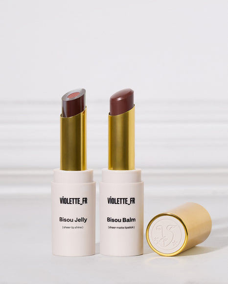 Two lipsticks in white and gold tubes are standing upright next to one another on a white surface. The left is a two-toned sheer purple shade, and the right is a matte cool brown. A cap rests nearby.