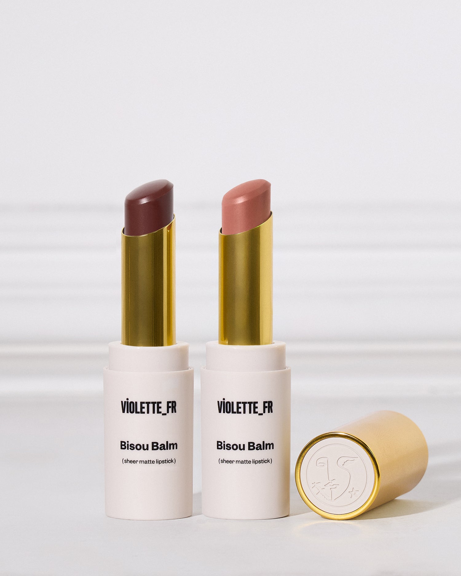 Two open Violette_Fr Bisou Balm lipstick tubes with gold caps, one showing a matte cool brown shade and the other a nude pink shade, with the gold cap lying beside them on a light gray background.