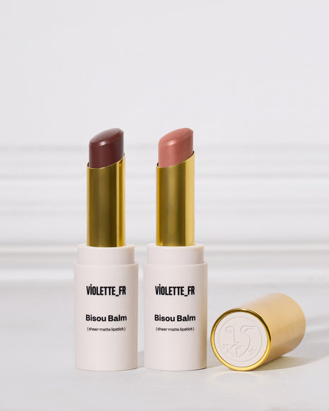 Two open Violette_Fr Bisou Balm lipstick tubes with gold caps, one showing a matte cool brown shade and the other a nude pink shade, with the gold cap lying beside them on a light gray background.