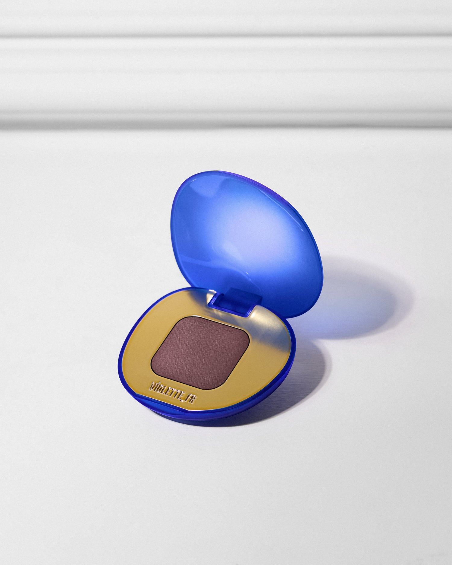 VIOLETTE_FR is embossed on the gold interior of an open, royal blue and gold compact, revealing a pan of matte deep plum eyeshadow.