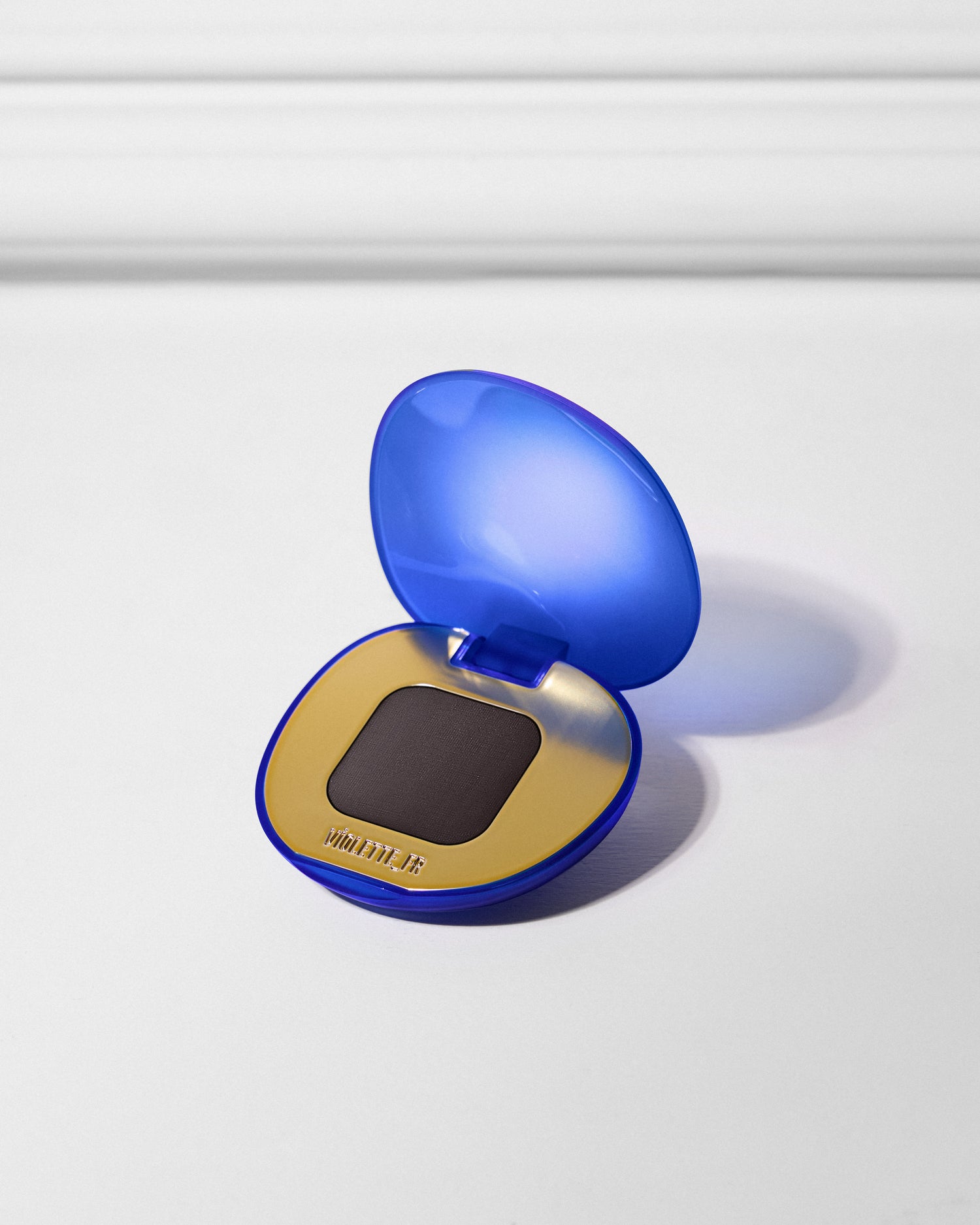 An open Violette_FR makeup compact with a translucent royal blue casing, gold interior, and a pan of dark matte brown/black powder.