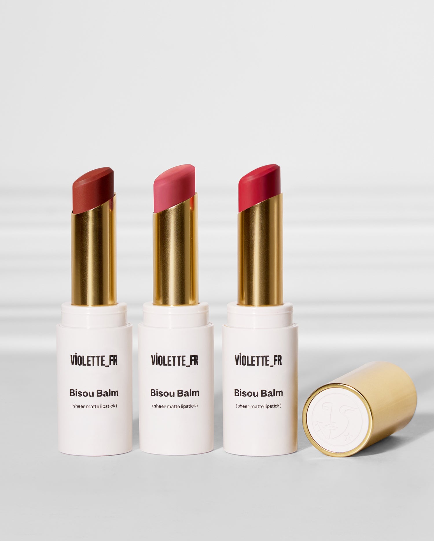 Bisou Balm Trio - Three 'Bisou Balm (sheer matte lipstick) VIOLETTE_FR' tubes, in reddish-brown, pink, and red, stand upright. A fourth gold-capped tube lies on its side. - Image 1 of 20