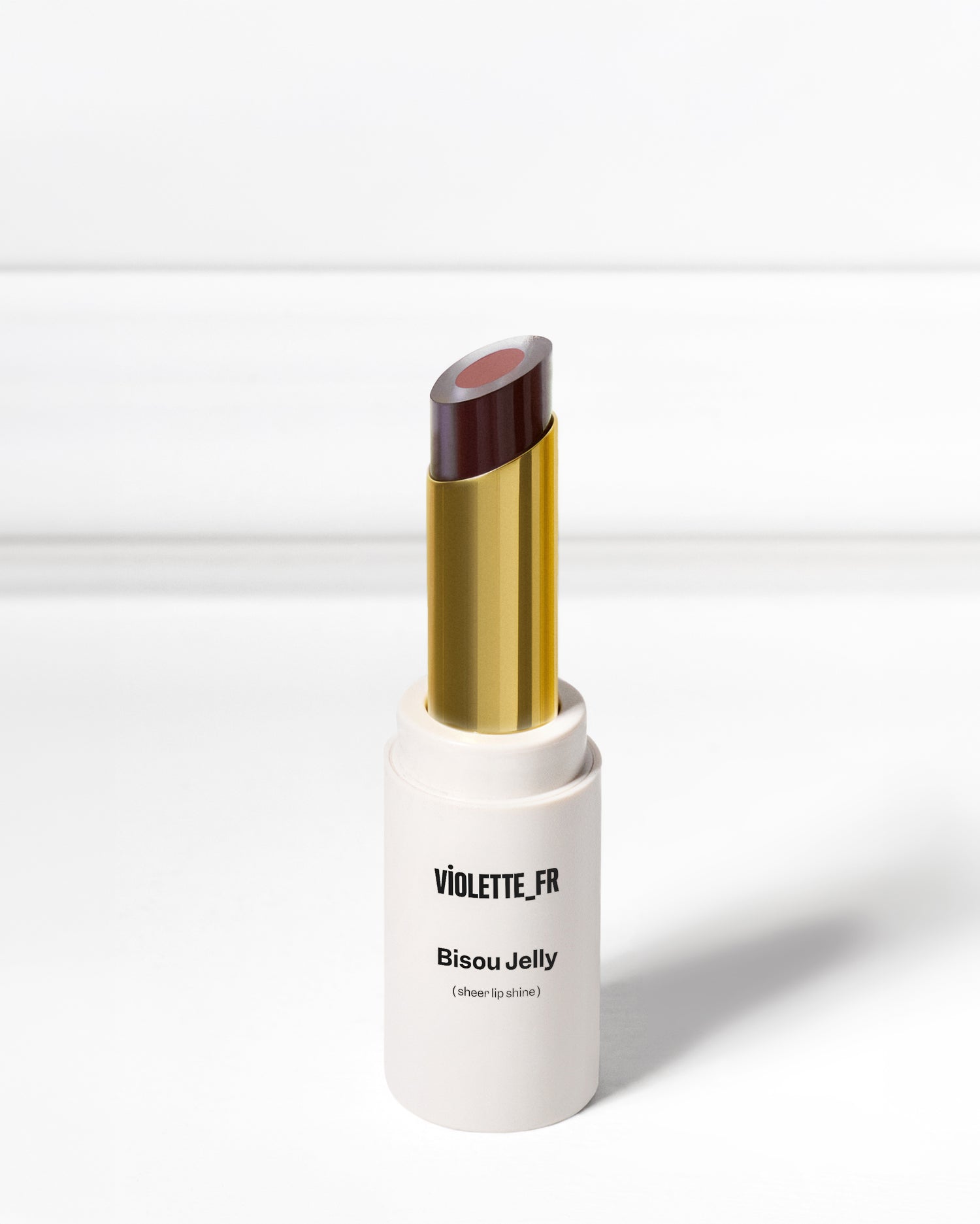 A white and gold tube of lipstick with a two-toned colored bullet stands upright on a gray surface, casting a shadow beneath it.