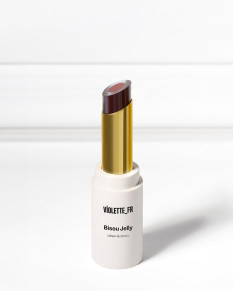 A white and gold tube of lipstick with a two-toned colored bullet stands upright on a gray surface, casting a shadow beneath it.