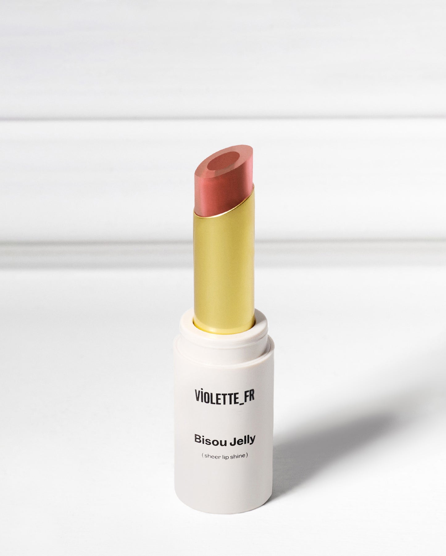 Bisou Jelly (sheer lip shine) by VIOLETTE_FR, an open tube of reddish-pink/coral lipstick with a gold inner casing, against a white background.