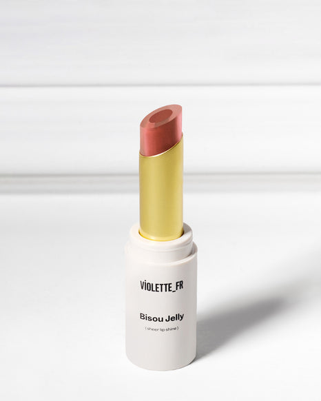 Bisou Jelly (sheer lip shine) by VIOLETTE_FR, an open tube of reddish-pink/coral lipstick with a gold inner casing, against a white background.