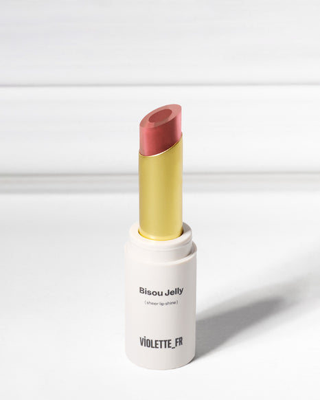 Bisou Jelly (sheer lip shine) by VIOLETTE_FR, an open tube of reddish-pink/coral lipstick with a gold inner casing, against a white background.