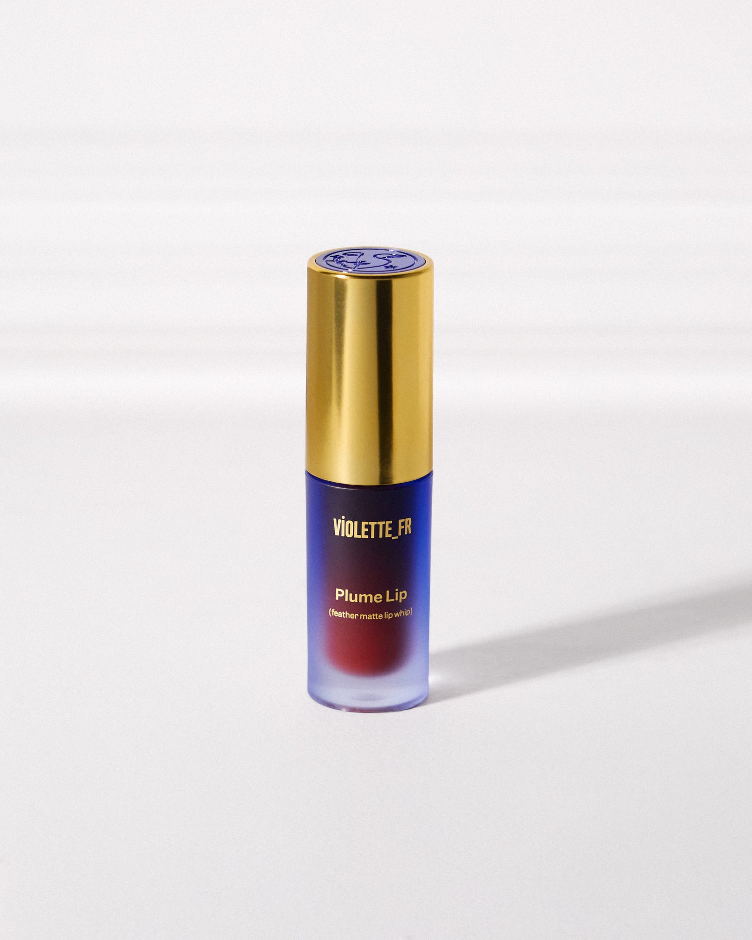 Violette_FR Plume Lip (feather-matte lip whip) in a frosted blue bottle with a gold cap, showing the deep red/burgundy product shade.