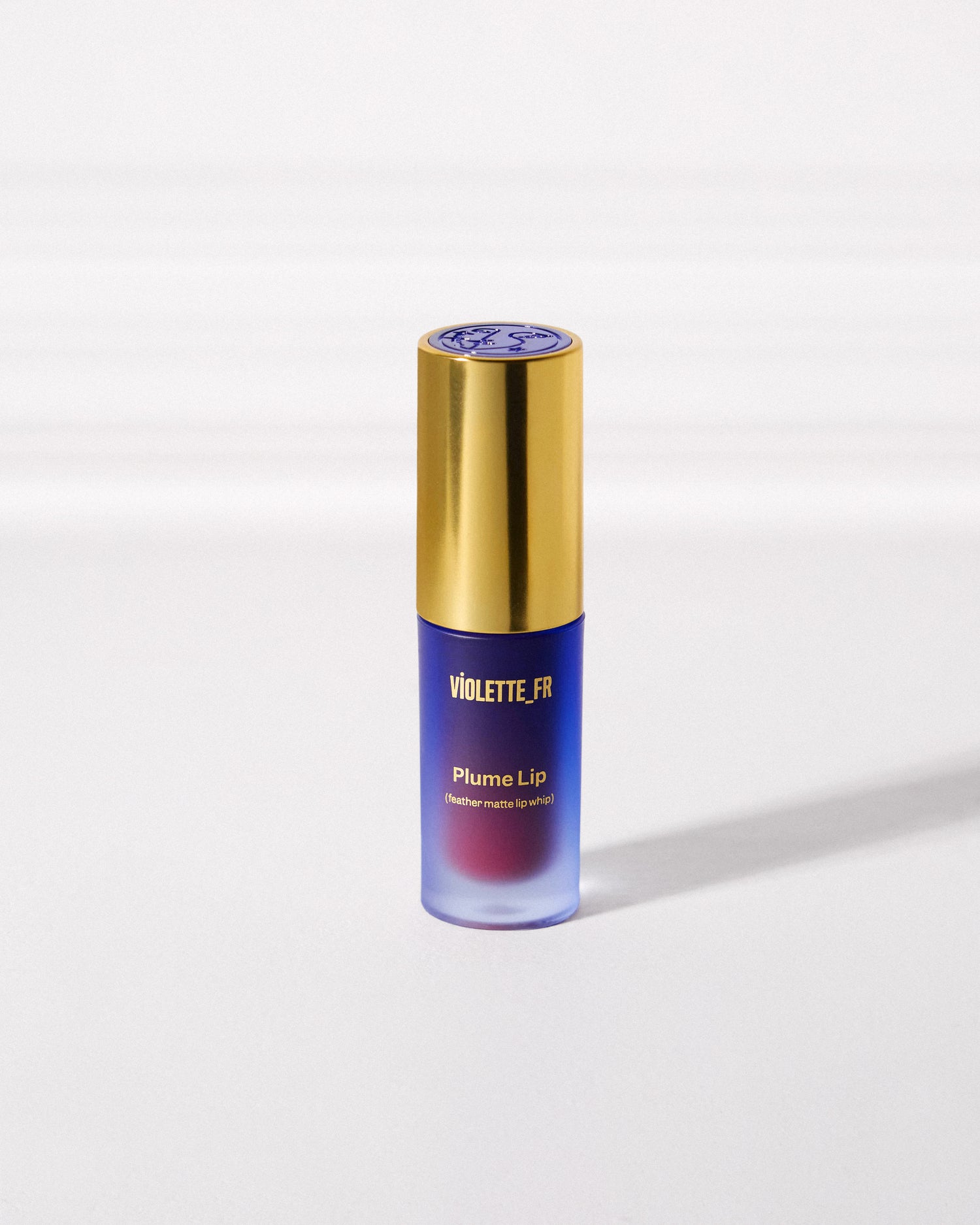VIOLETTE_FR Plume Lip (feather matte lip whip) in a blue frosted bottle with a gold cap, showing the reddish-pink product inside.