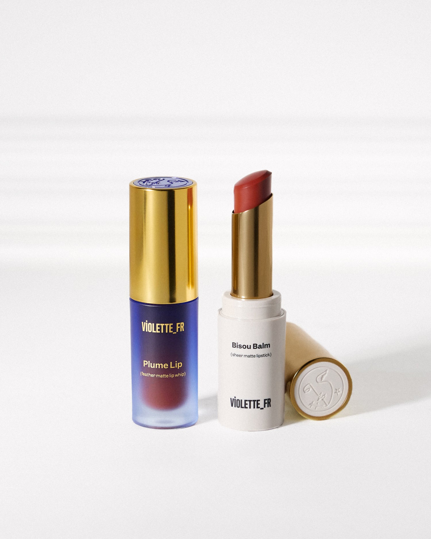 Violette_FR Plume Lip (feather matte lip whip) in a purple-to-red tube and Bisou Balm (sheer matte lipstick) with a reddish-brown bullet in a white tube, both with gold accents.