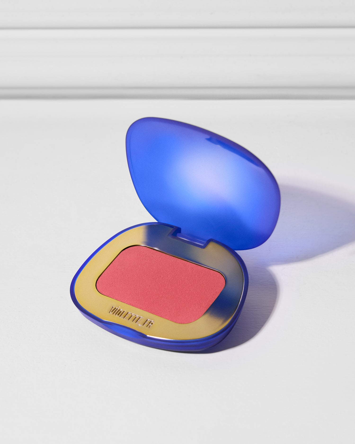 VIOLETTE_FR is embossed on the gold rim of an open, blue compact, revealing a coral-pink powder.