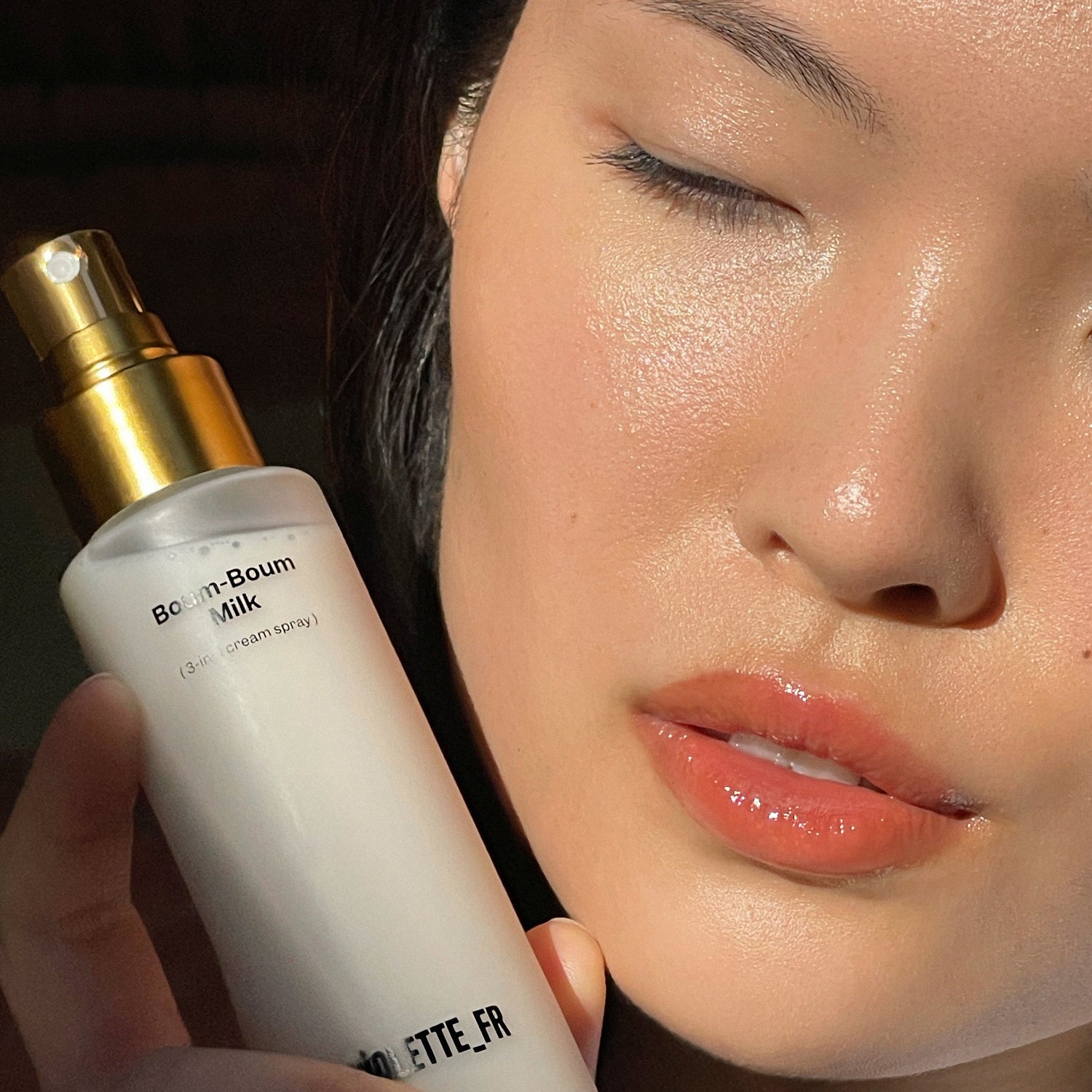 Up close model holding skincare bottle with gold sprayer top and a glossy lip
