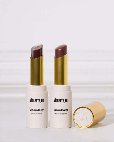 Two lipsticks in white and gold tubes are standing upright next to one another on a white surface. The left is a two-toned sheer purple shade, and the right is a matte cool brown. A cap rests nearby.