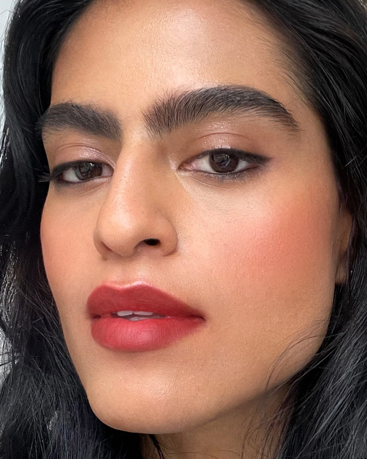 Woman with medium skin tone wearing a bright red diffused lipstick on her lips. There is thin eyeliner on her eyes and a slight shimmer on her eyelids. 