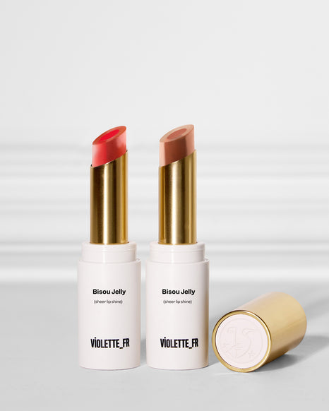 Two Violette_FR Bisou Jelly sheer lip shine lipsticks, one coral and one nude, stand upright. A gold cap with an embossed logo lies beside them.