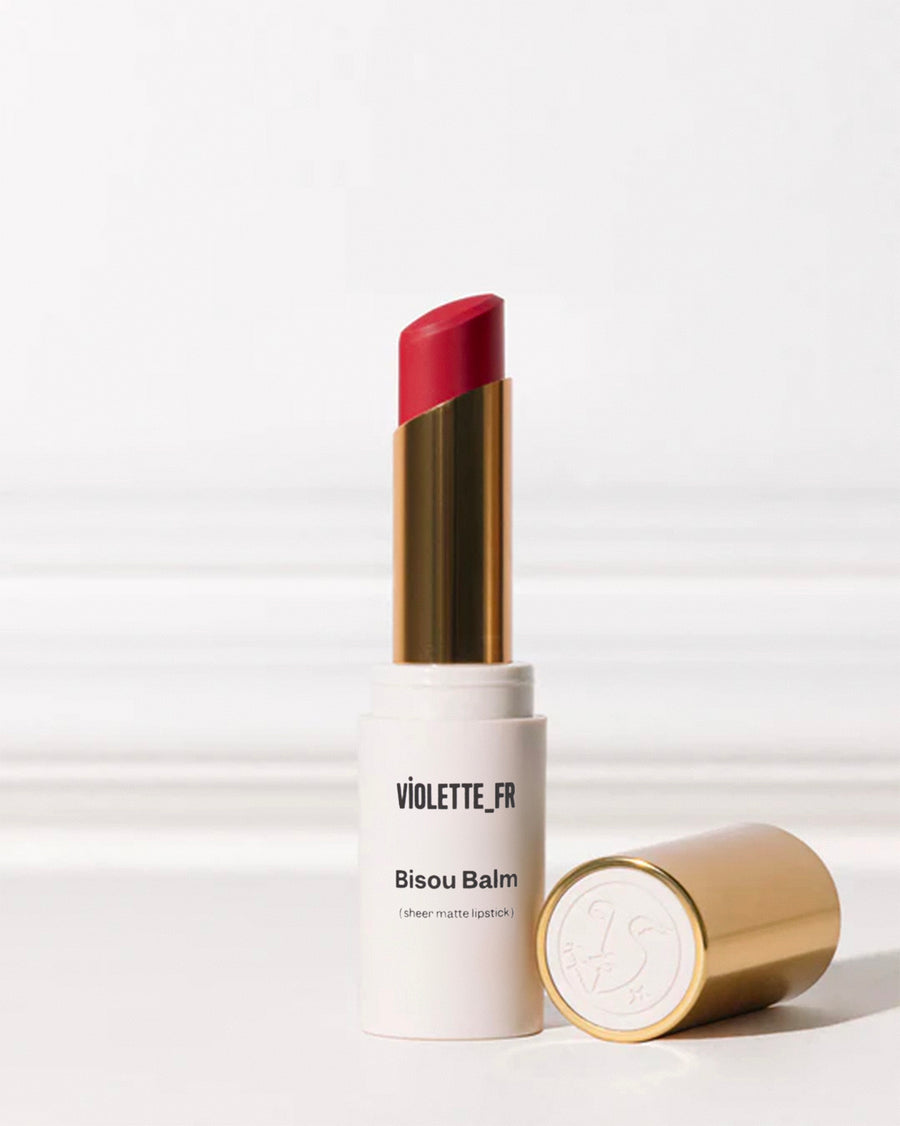 Bisou Balm (sheer matte lipstick) by VIOLETTE_FR: a matte off-white tube with a vibrant red bullet extended, next to its gold cap with an embossed logo.