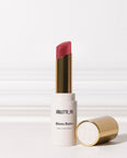 Bisou Balm (sheer matte lipstick) by VIOLETTE_FR. A white lipstick tube with gold accents stands upright, revealing a reddish-pink lipstick bullet, with its gold cap lying beside it.
