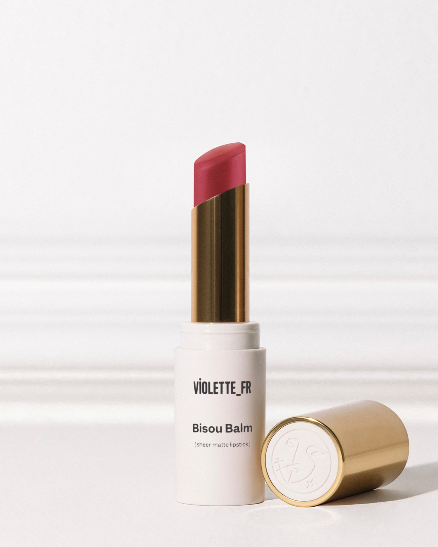 Bisou Balm - Bêtise - Bisou Balm (sheer matte lipstick) by VIOLETTE_FR. A white lipstick tube with gold accents stands upright, revealing a reddish-pink lipstick bullet, with its gold cap lying beside it. - Image 41 of 41