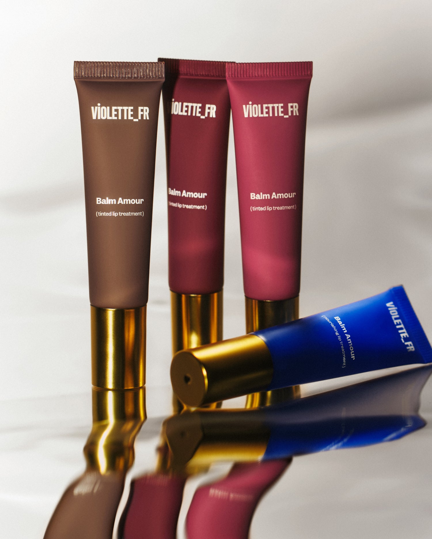 Three tubes of lip balm in different shades with gold caps stand upright on a reflective surface. A blue tube of the lip balm lies in the foreground. 