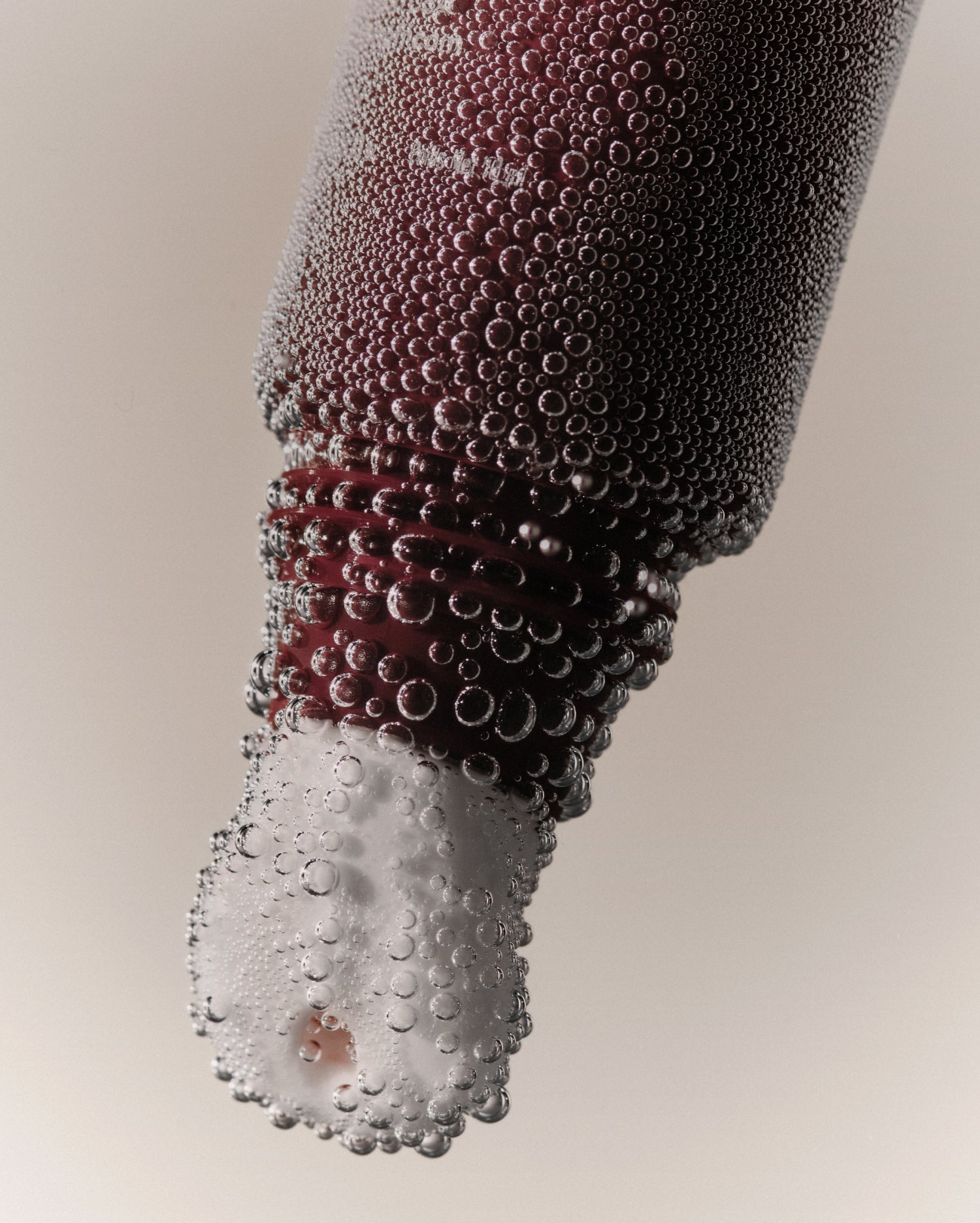 A close-up of an opened tube of maroon lip balm with a white applicator facing down. The tube is covered in condensation droplets against a neutral background. 