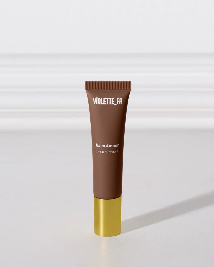 A brown tube of lip balm with a gold cap stands upright on a gray surface, casting a shadow beneath it. 