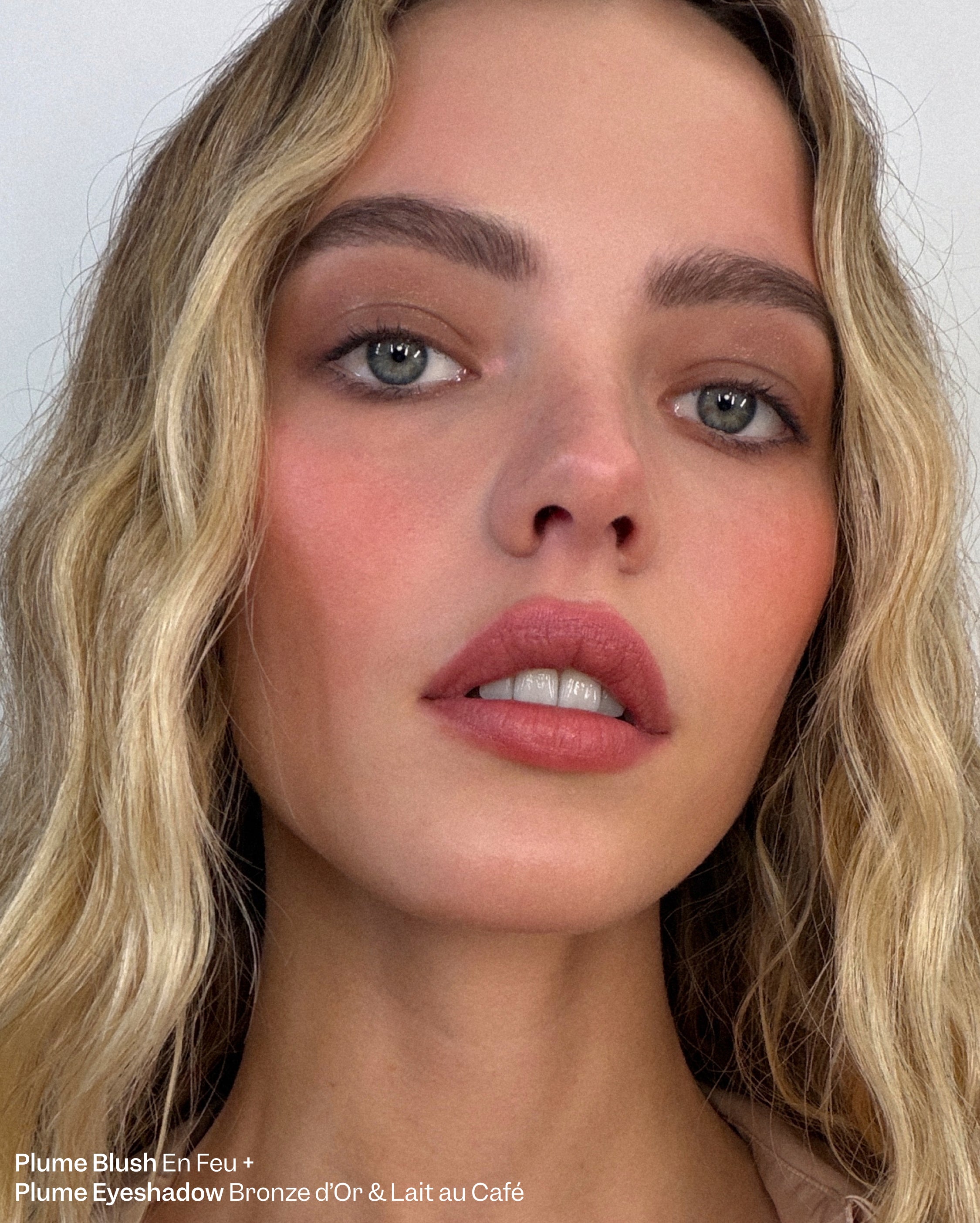Plume Blush En Feu + Plume Eyeshadow Bronze d'Or & Lait au Café. A woman with light skin, blonde hair, and green eyes wears a warm peachy-pink blush, subtle bronze eyeshadow, and muted pink lipstick.