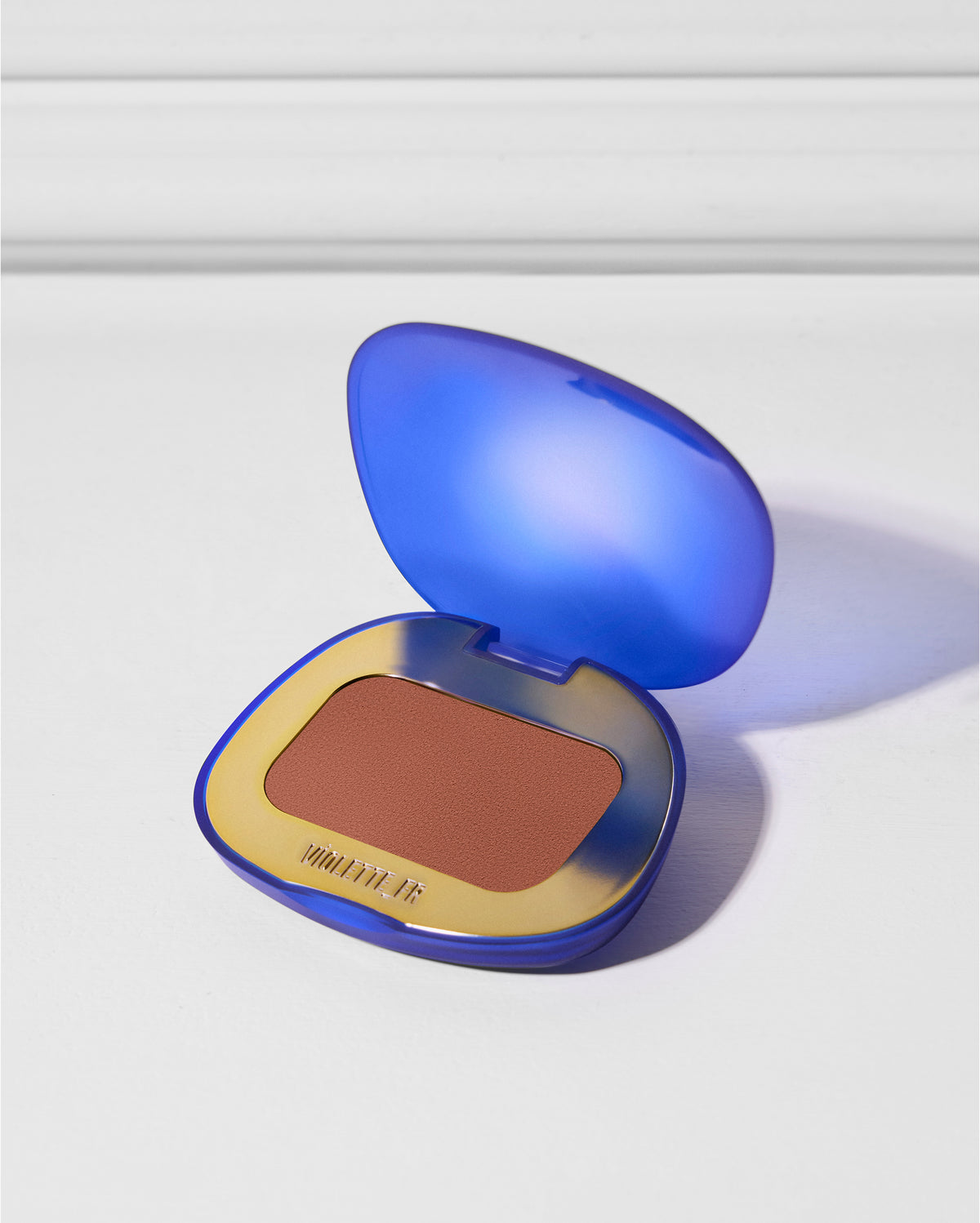 Open compact with a translucent blue lid and gold base, revealing a matte reddish-brown powder. The brand name VIOLETTE_FR is visible on the base.