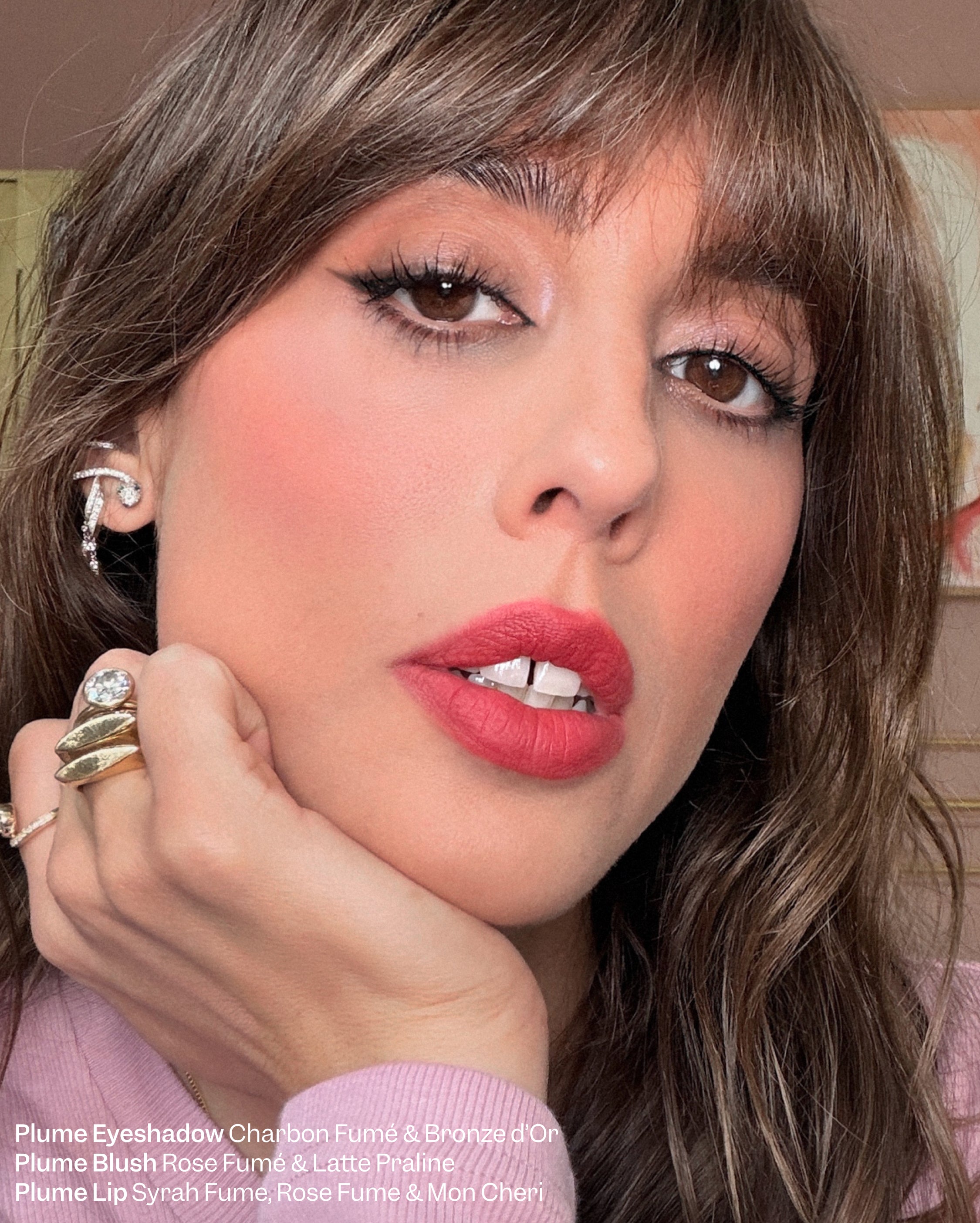 A woman with light to medium skin and brown hair wears a pink toned lipstick and blush on cheeks with chin resting on hand
