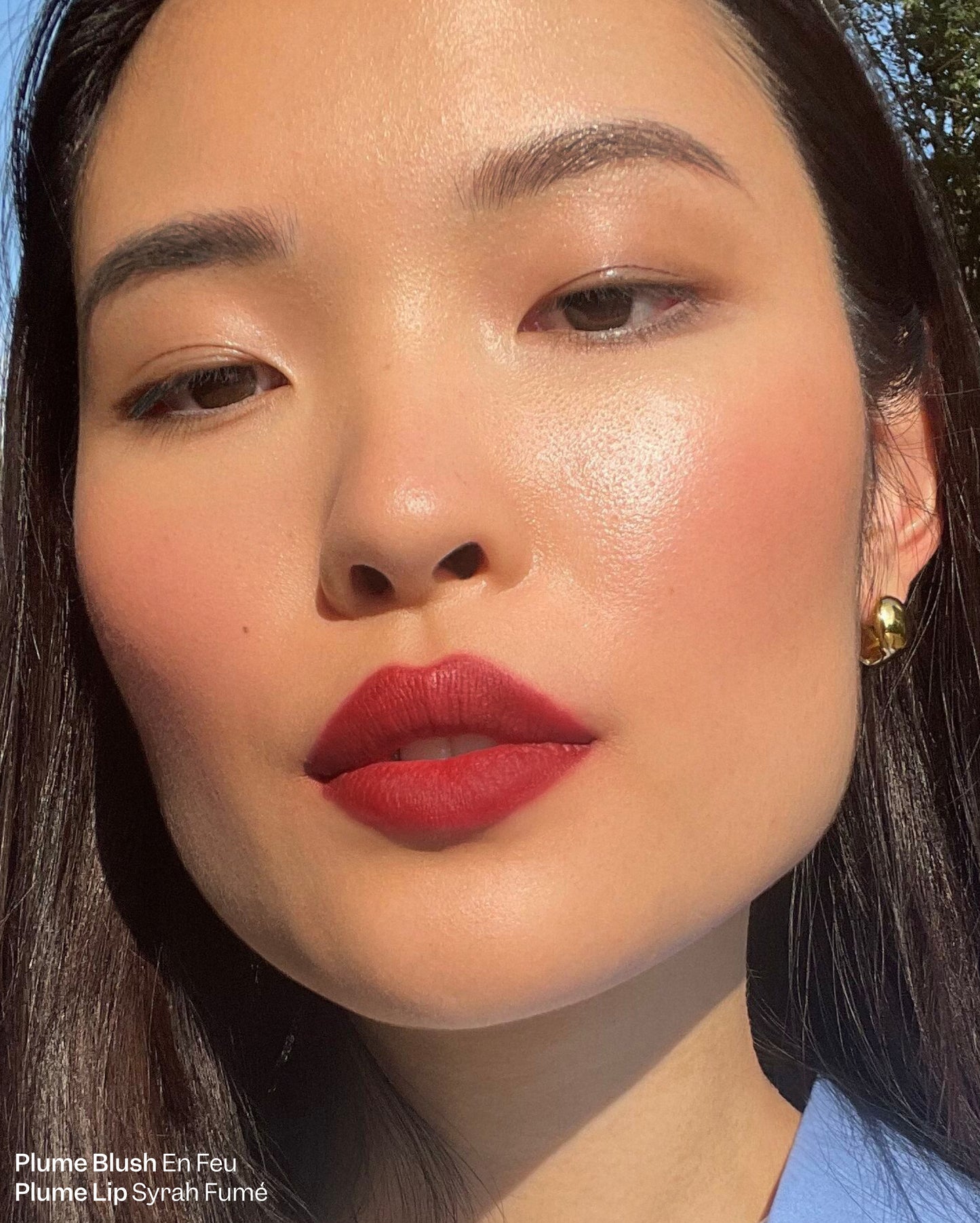 Plume Blush En Feu and Plume Lip Syrah Fume. Up close shot of woman with dark brown hair wearing bold red lipstick and blush - Plume Essentials Trio