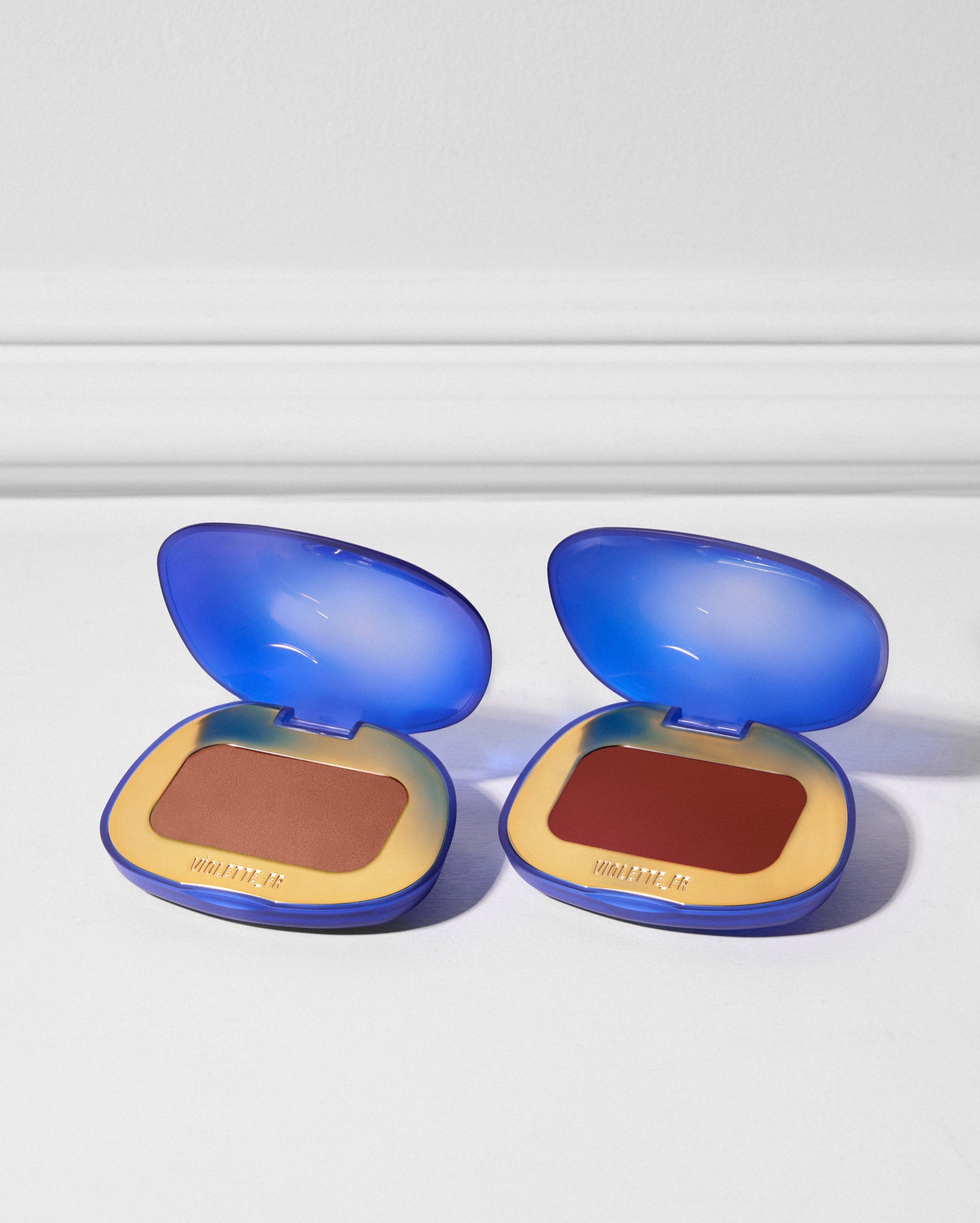 Two open blue compacts with gold interiors, each embossed with "VIOLETTE_FR". The left compact contains a reddish-brown shade, and the right contains a deep red shade.