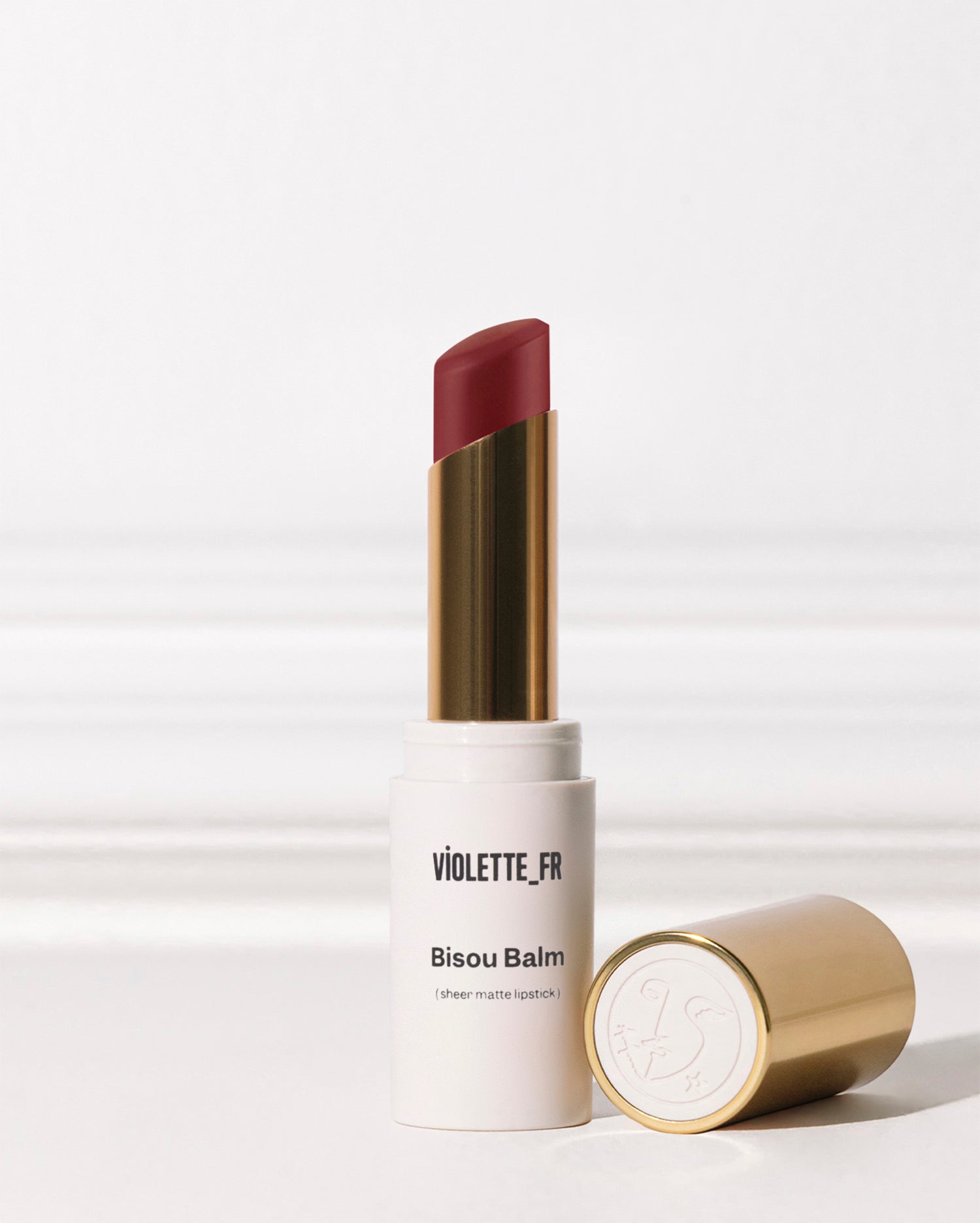 Bisou Balm - Bêtise - Violette_FR Bisou Balm (sheer matte lipstick) in a deep red shade, with its gold cap featuring a stylized face logo lying beside it. - Image 62 of 62