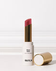 Bisou Balm (sheer matte lipstick) by VIOLETTE_FR. A white lipstick tube with gold accents stands upright, revealing a reddish-pink lipstick bullet, with its gold cap lying beside it.
