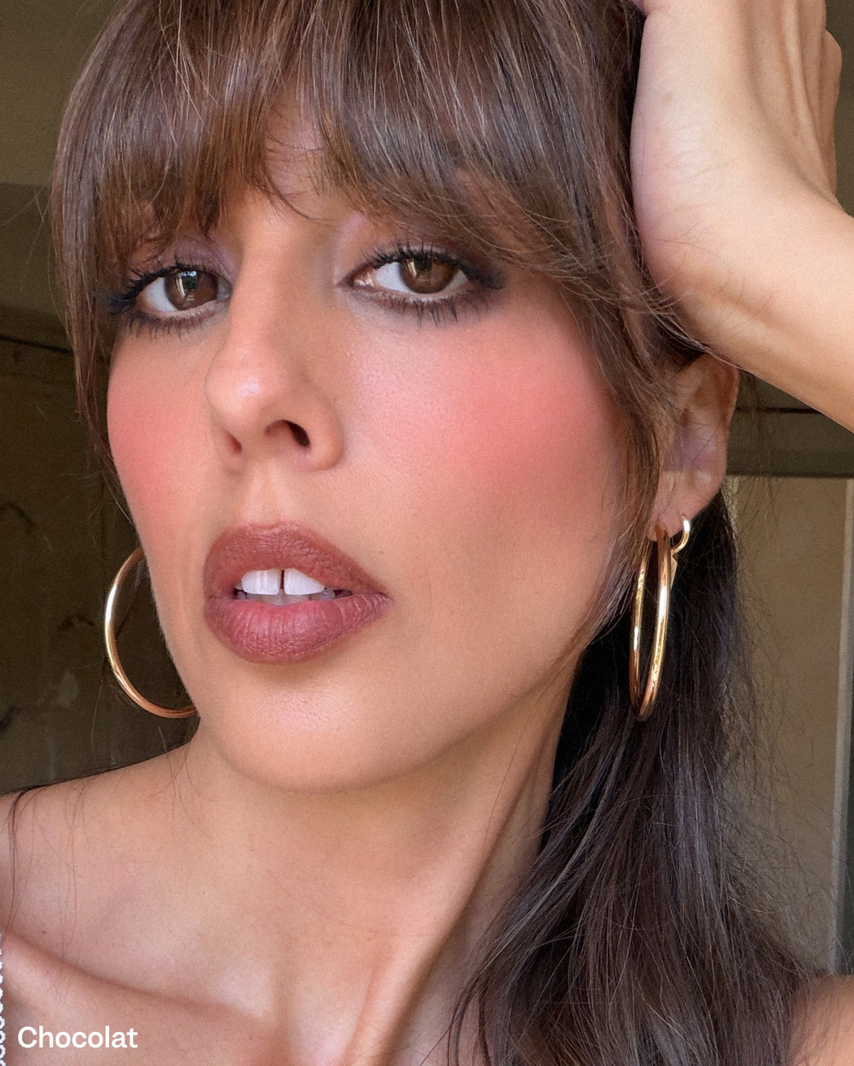 Bisou Balm - Calisson - Chocolat: Close-up of a woman with medium skin tone, brown hair, and bangs, wearing cool brown with burgundy undertones lipstick and subtle eye makeup, with two gold hoop earrings. - Image 24 of 24