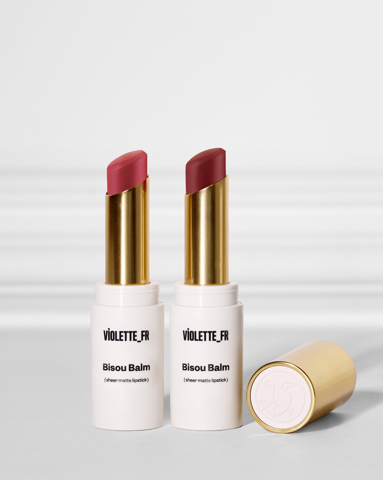 Two Violette_FR Bisou Balm sheer matte lipsticks, one in muted dark mauve pink and one in dark reddish brown, with a gold cap showing the embossed logo.
