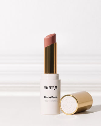 Bisou Balm (sheer matte lipstick) by VIOLETTE_FR, shown in an open white and gold tube with a muted pinkish-brown shade, with its gold cap lying next to it.