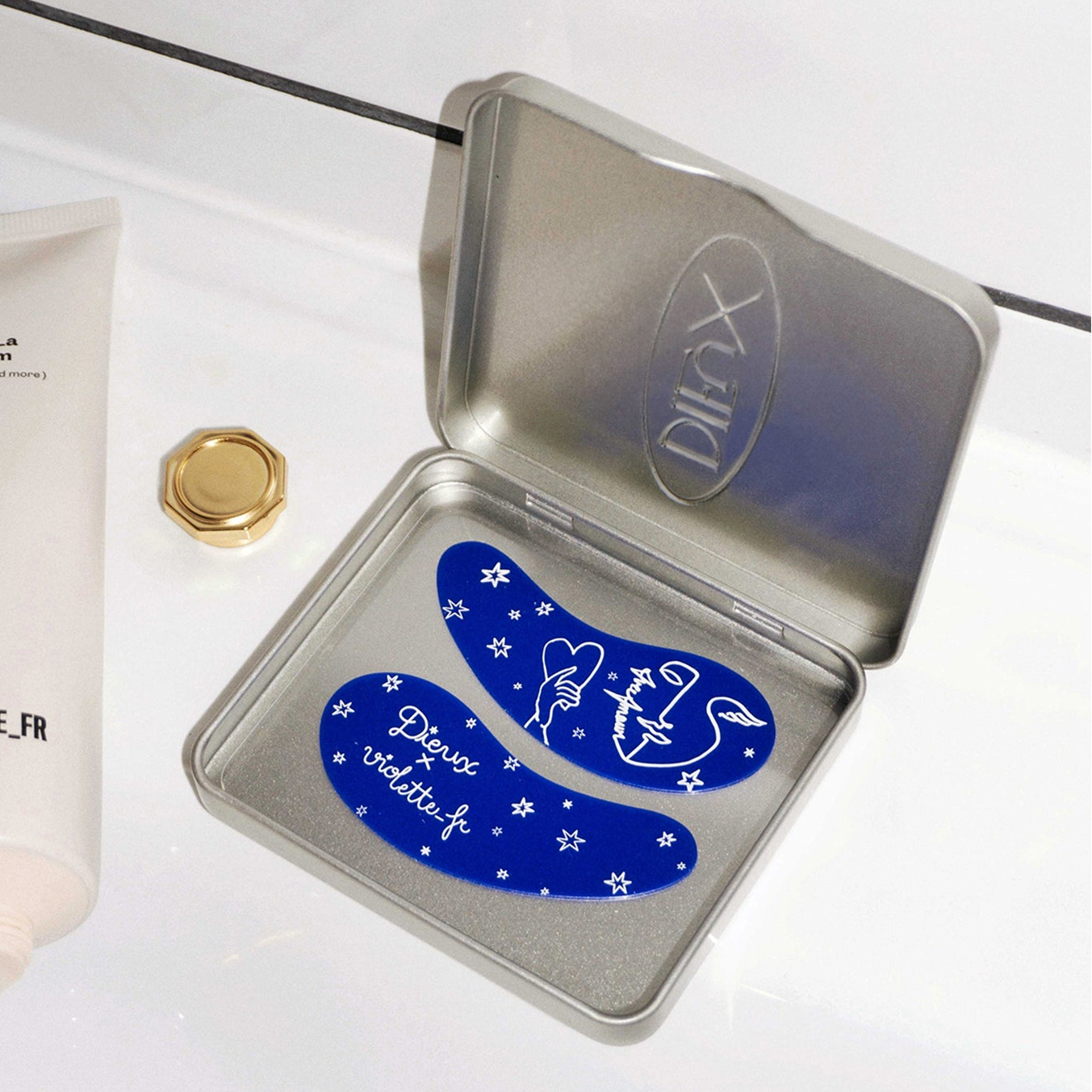Violette_FR C'est La Cream tube and a gold cap next to an open silver Dieux tin containing two blue eye patches. The patches feature white designs of stars, a hand holding a heart, a face, a swan-like figure, and the text 'Dieux violette.fr' and 'Faire l'Amour'.